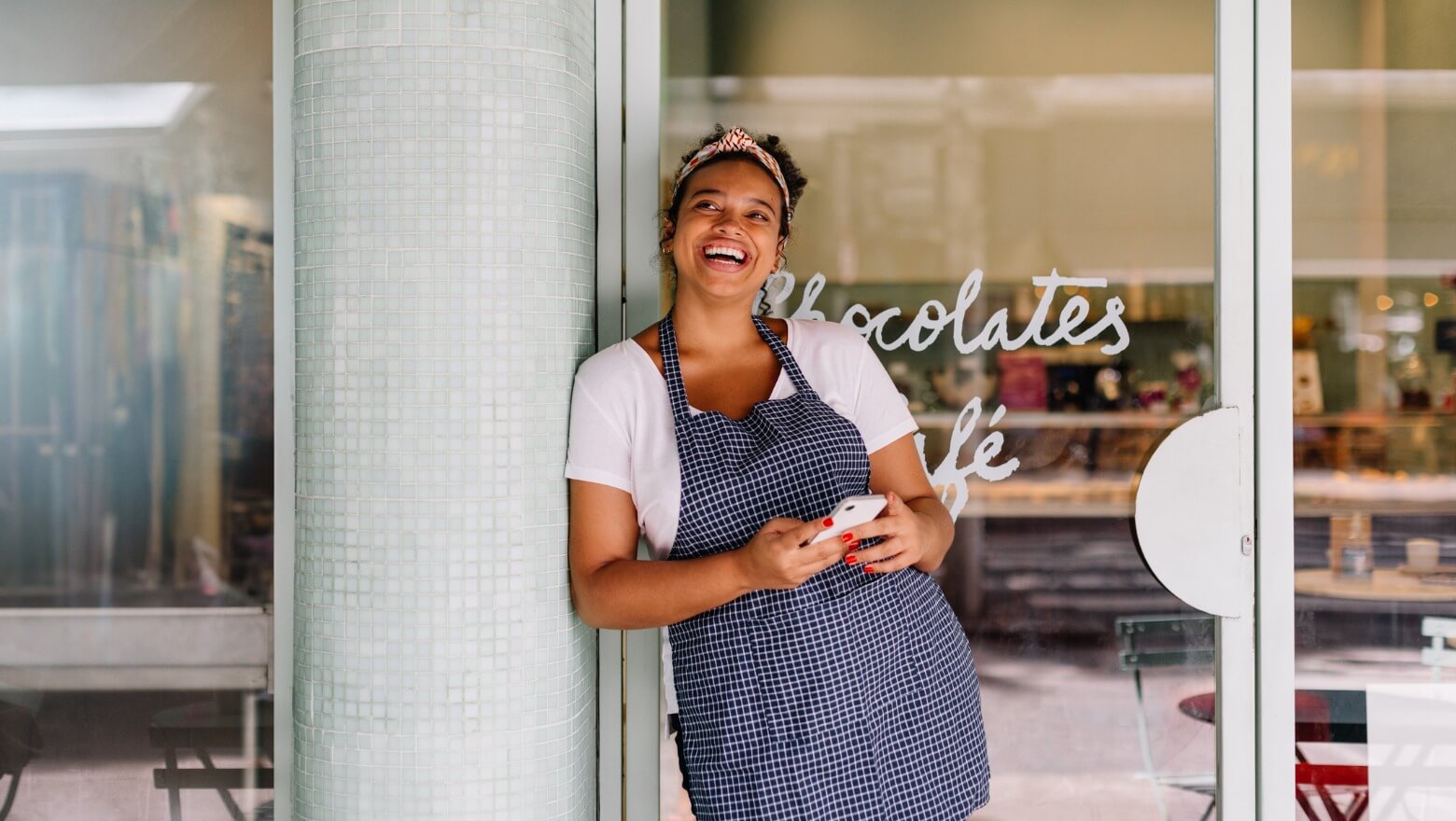 A cheerful woman in a checkered apron is standing outside a cafe, smiling and looking at her phone.