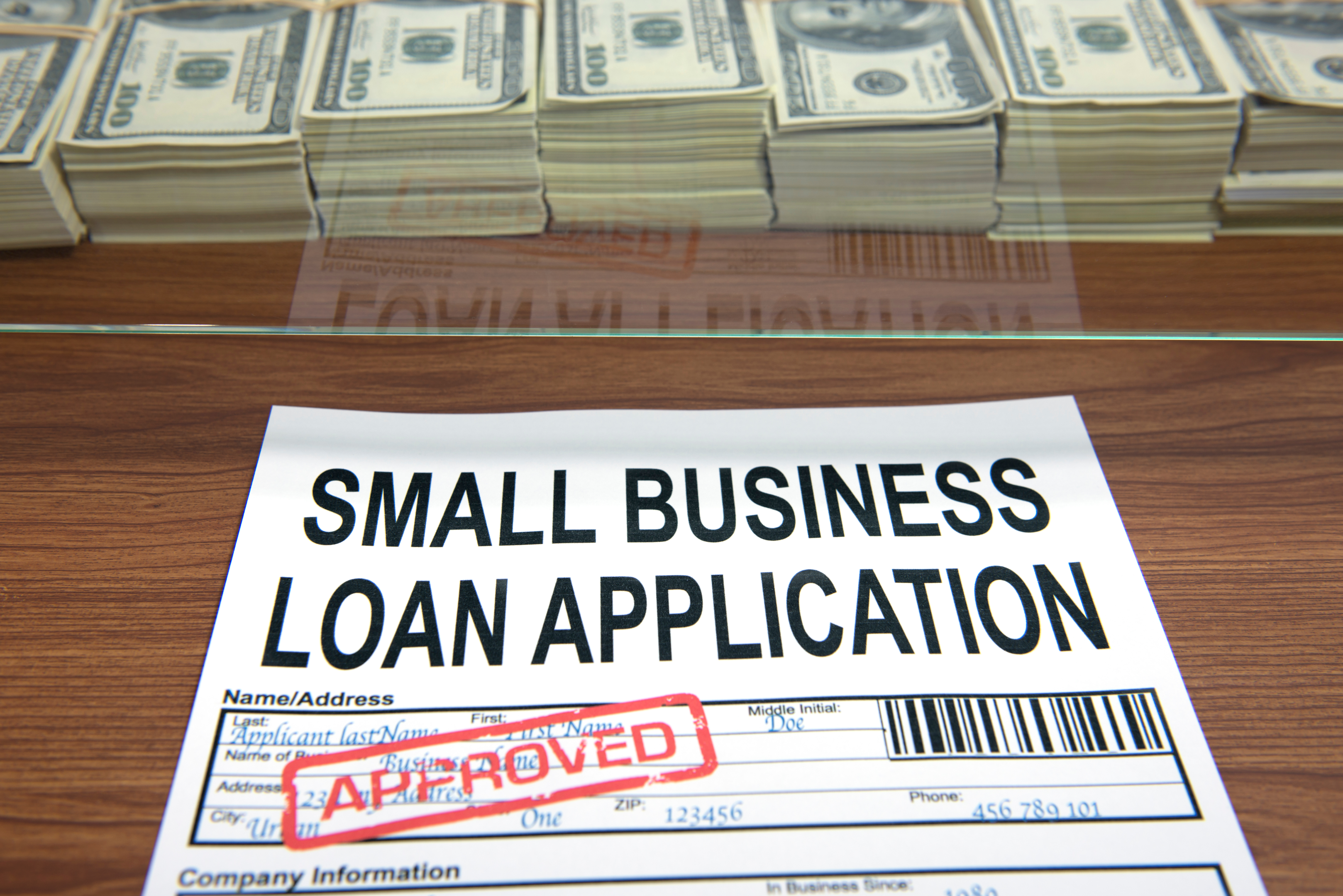 The image shows a small business loan application form with an "Approved" stamp, placed on a wooden surface with stacks of hundred-dollar bills in the background.