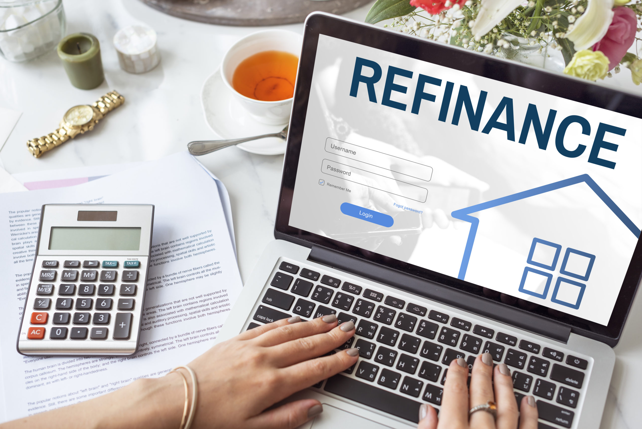 A person is working on a laptop with the word "REFINANCE" displayed on the screen, surrounded by documents, a calculator, a cup of tea, and a watch, suggesting financial planning or loan refinancing.