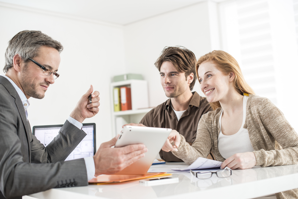 A man in a suit showing a tablet to a smiling couple during a discussion or consultation in an office setting.