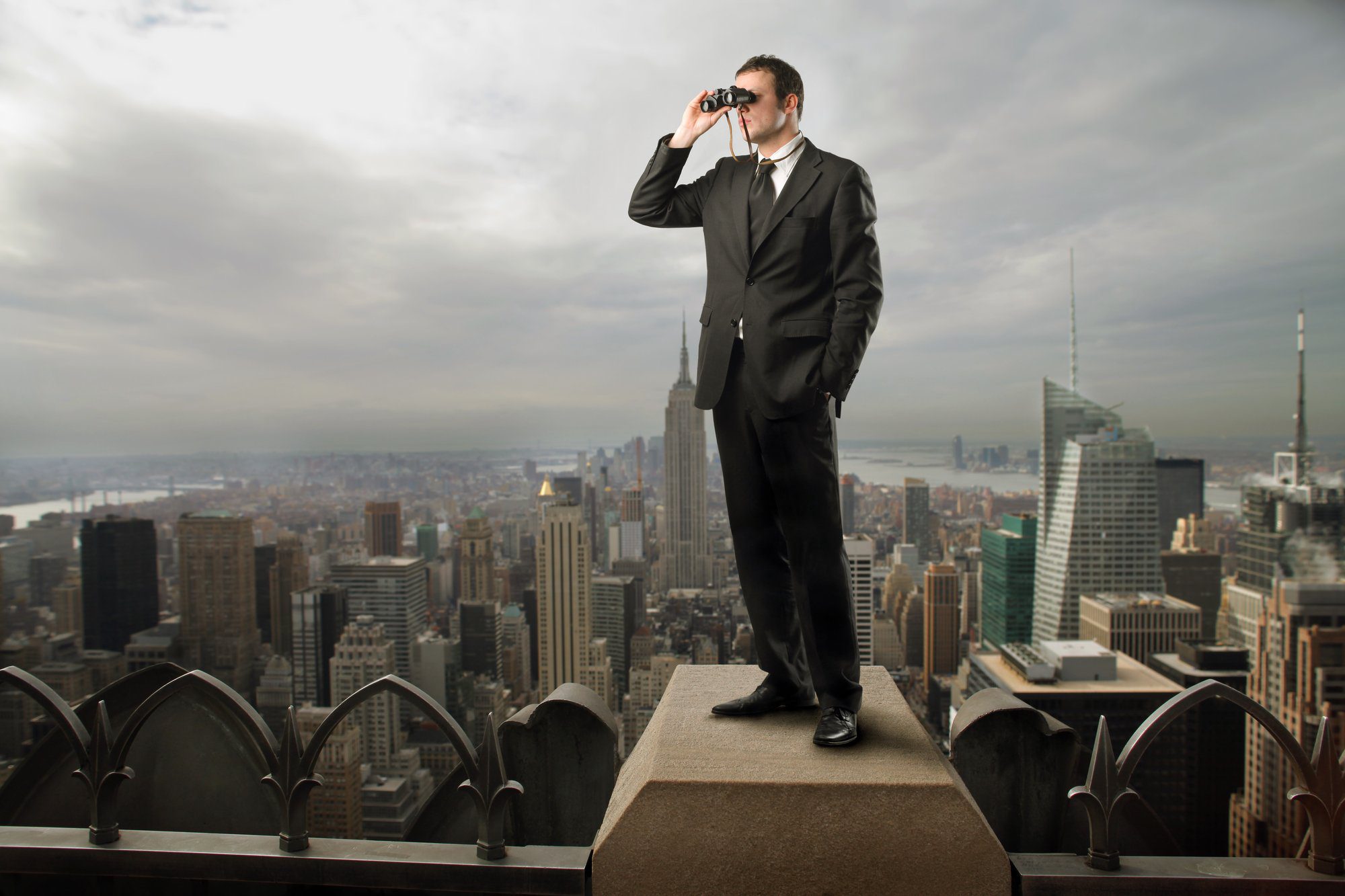 A man in a suit stands on a rooftop ledge overlooking a city skyline, using binoculars to observe the view.
