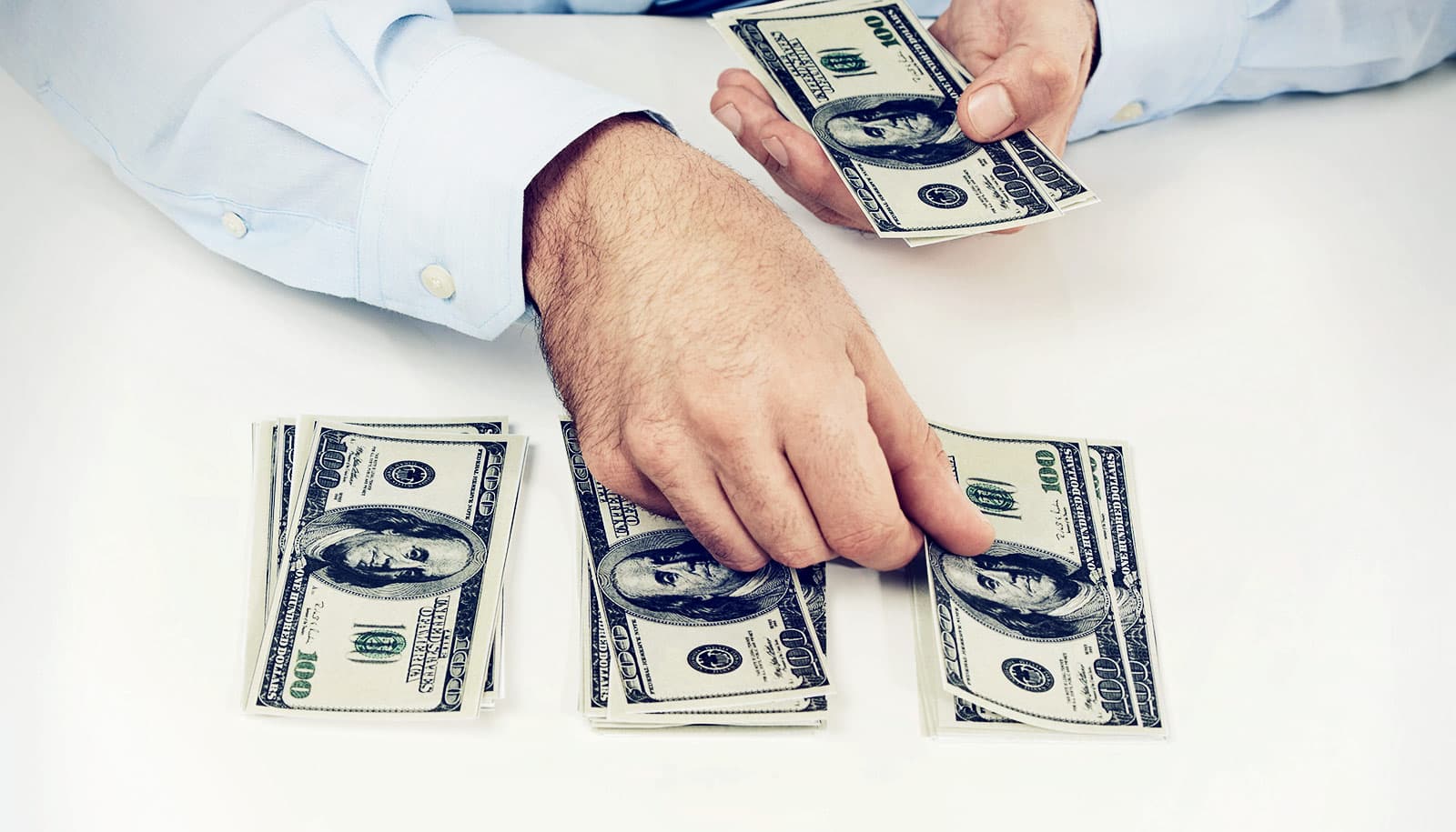 A person in a light blue shirt is counting and handling stacks of hundred-dollar bills on a white surface.
