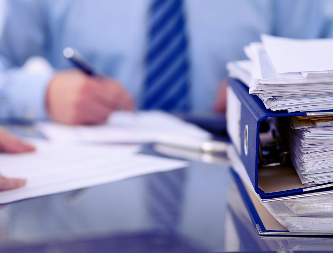 The image depicts a person in a business suit reviewing or filling out paperwork at a desk, with a large stack of documents or files nearby.
