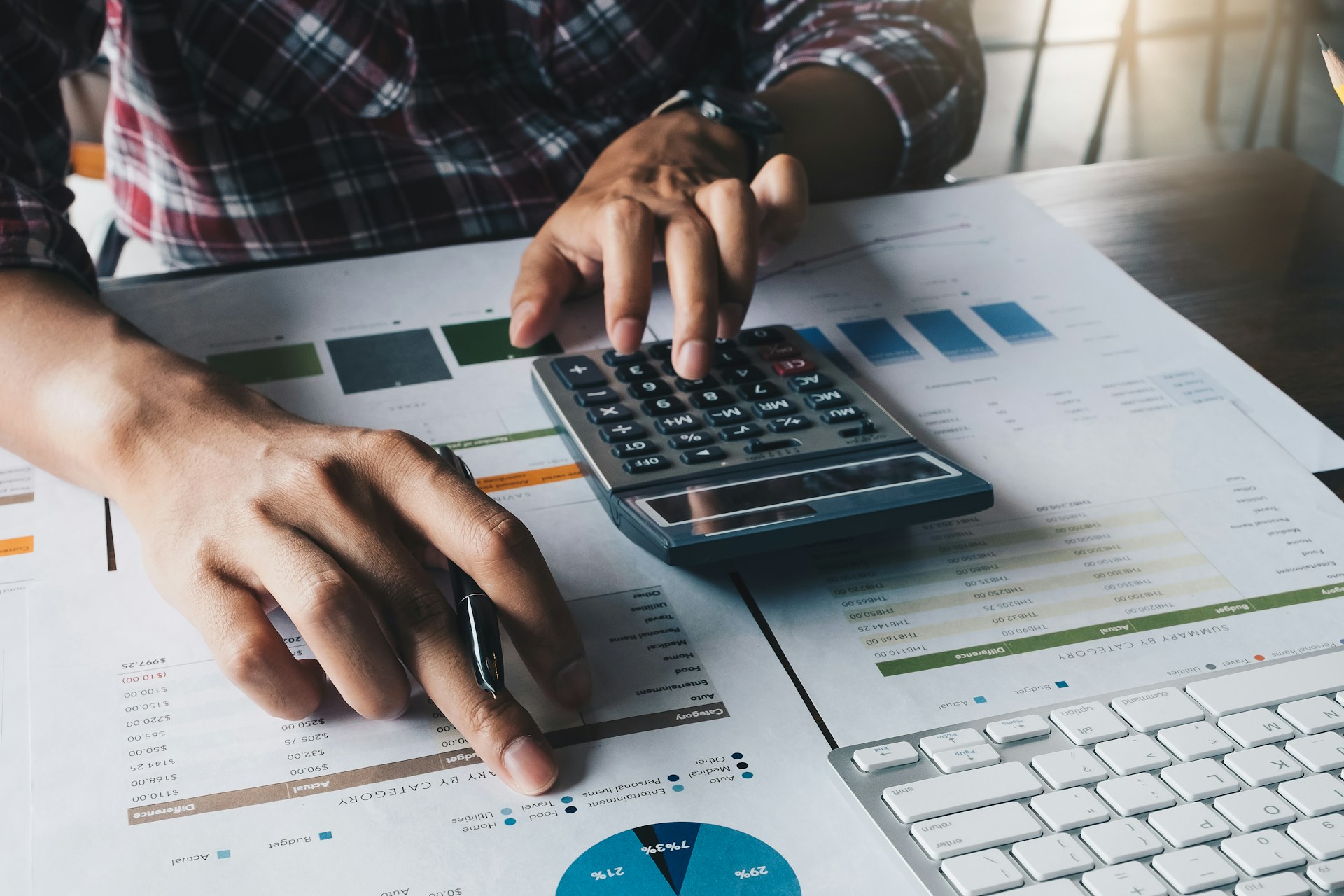 The image shows a person working on financial analysis or calculations, using a calculator, pen, and various documents on a desk.