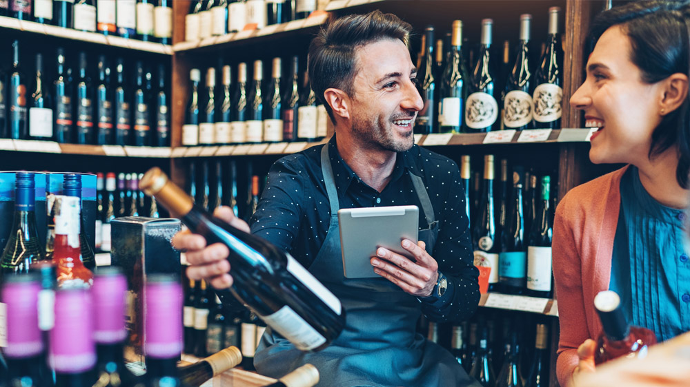 The image shows a man and a woman happily interacting in a wine store, with the man holding a bottle of wine and a tablet.