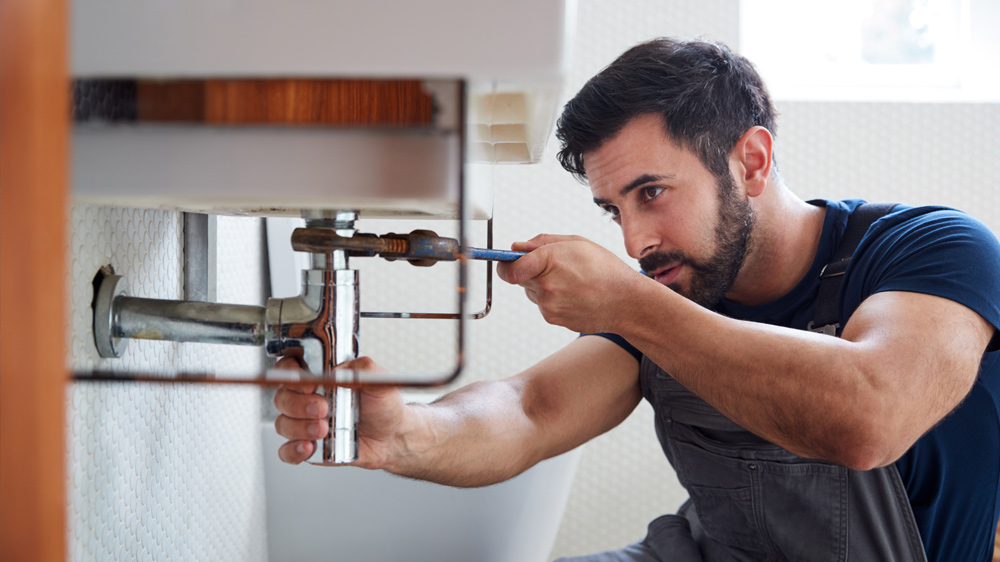 The image shows a man repairing or fixing plumbing under a sink.