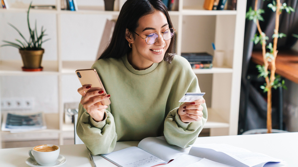 The image shows a woman happily looking at her phone and a credit card while sitting at a desk with a cup of coffee, an open notebook, and a book.