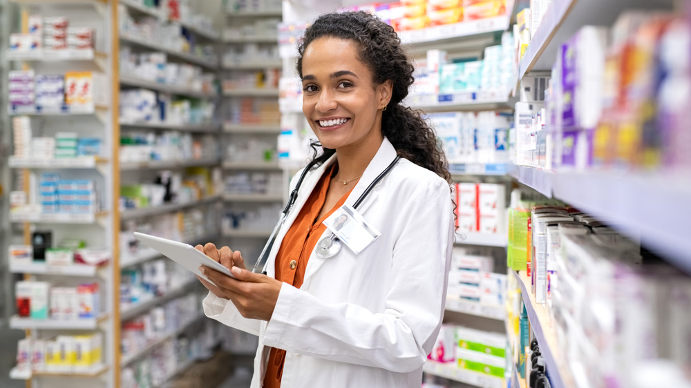 A smiling female healthcare professional or pharmacist is standing in a pharmacy aisle, holding a tablet.