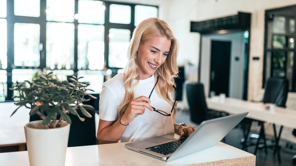A woman is smiling and working on a laptop in a modern, bright office space.