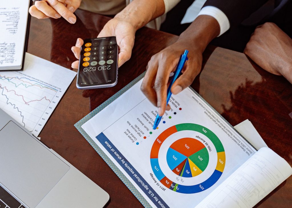 The image shows two individuals analyzing financial or business data, with one person holding a calculator and the other pointing at a colorful pie chart on a report.