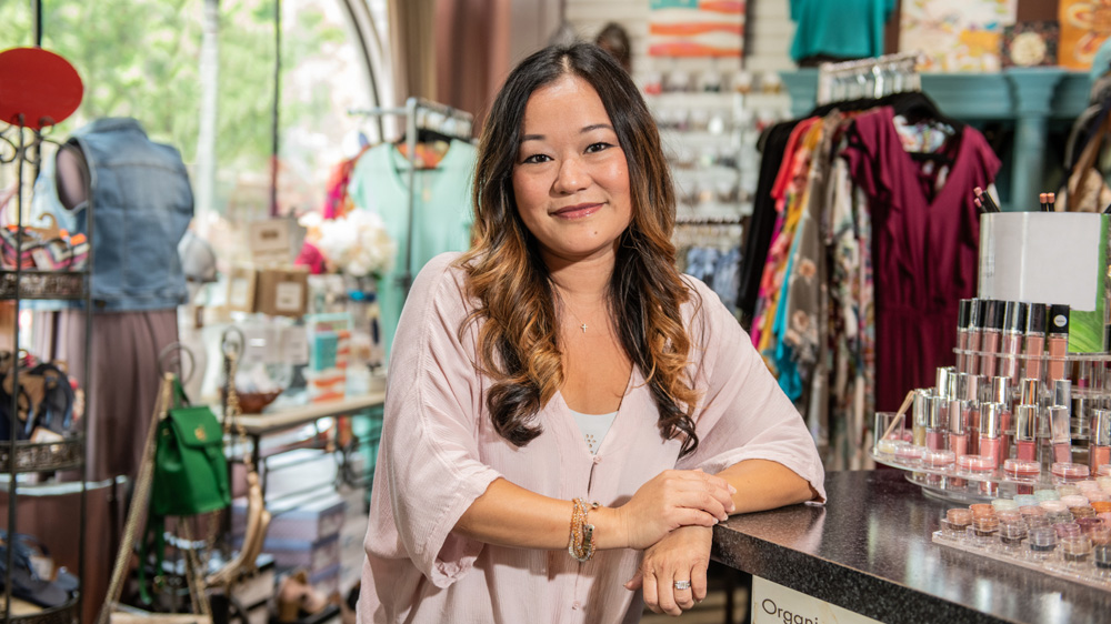 The woman is smiling confidently behind the counter of a boutique or beauty shop, surrounded by clothing and cosmetic products.