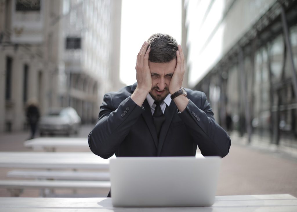 The man appears stressed and overwhelmed while working on his laptop outdoors in an urban setting.