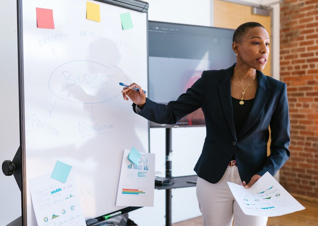 The woman is giving a presentation or explanation about budget planning in a professional setting.