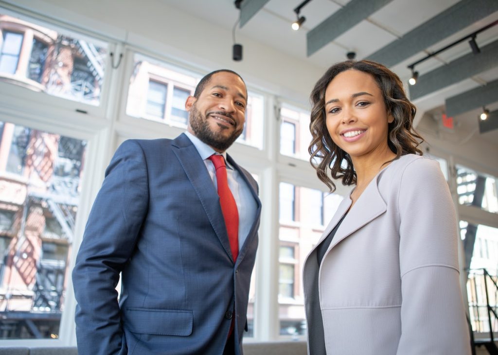 The picture shows two professionally dressed individuals smiling confidently in a modern office setting.