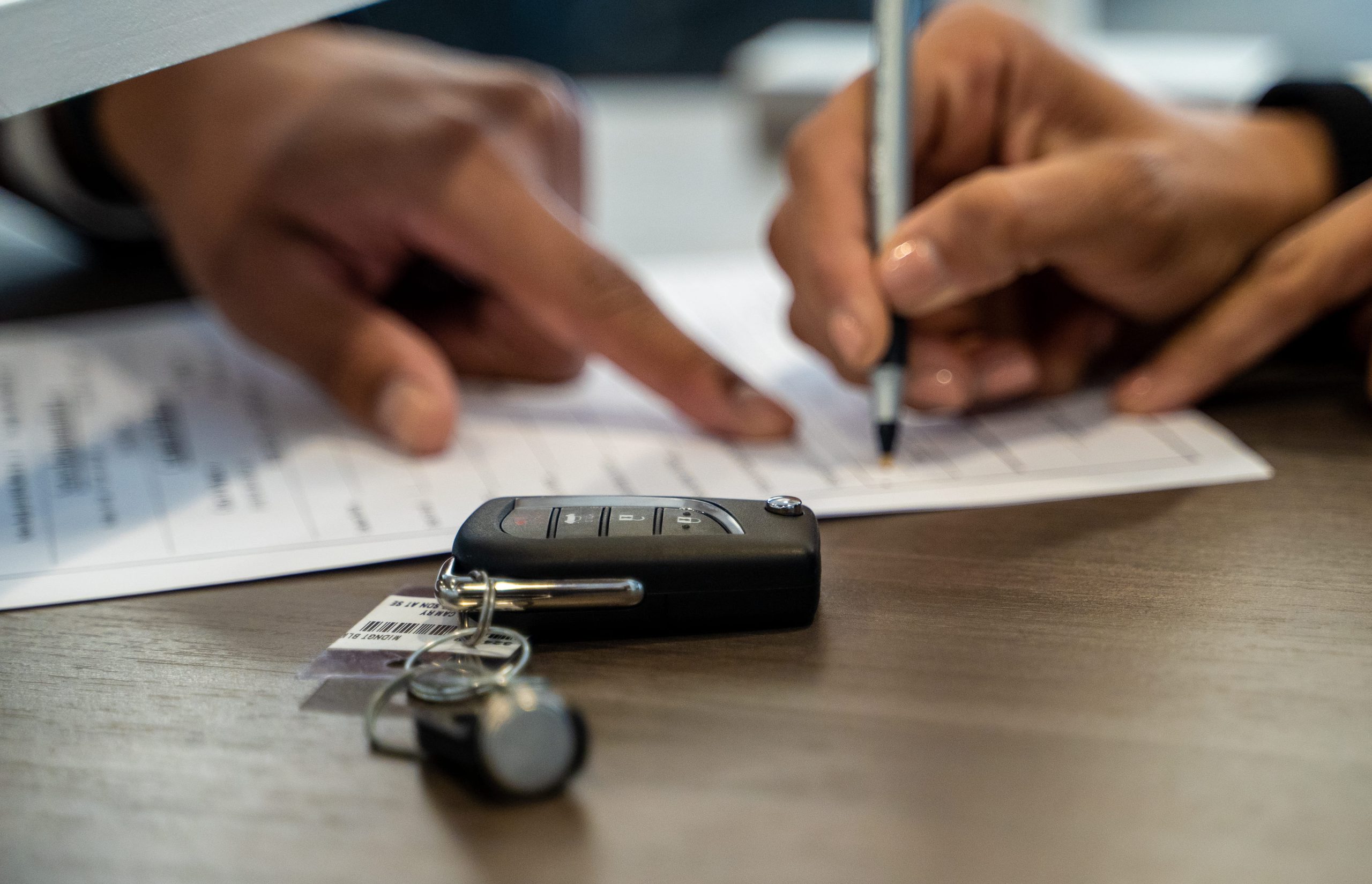 The image shows a person filling out a form or document, with a car key placed on the table, suggesting a process related to car ownership or financing.