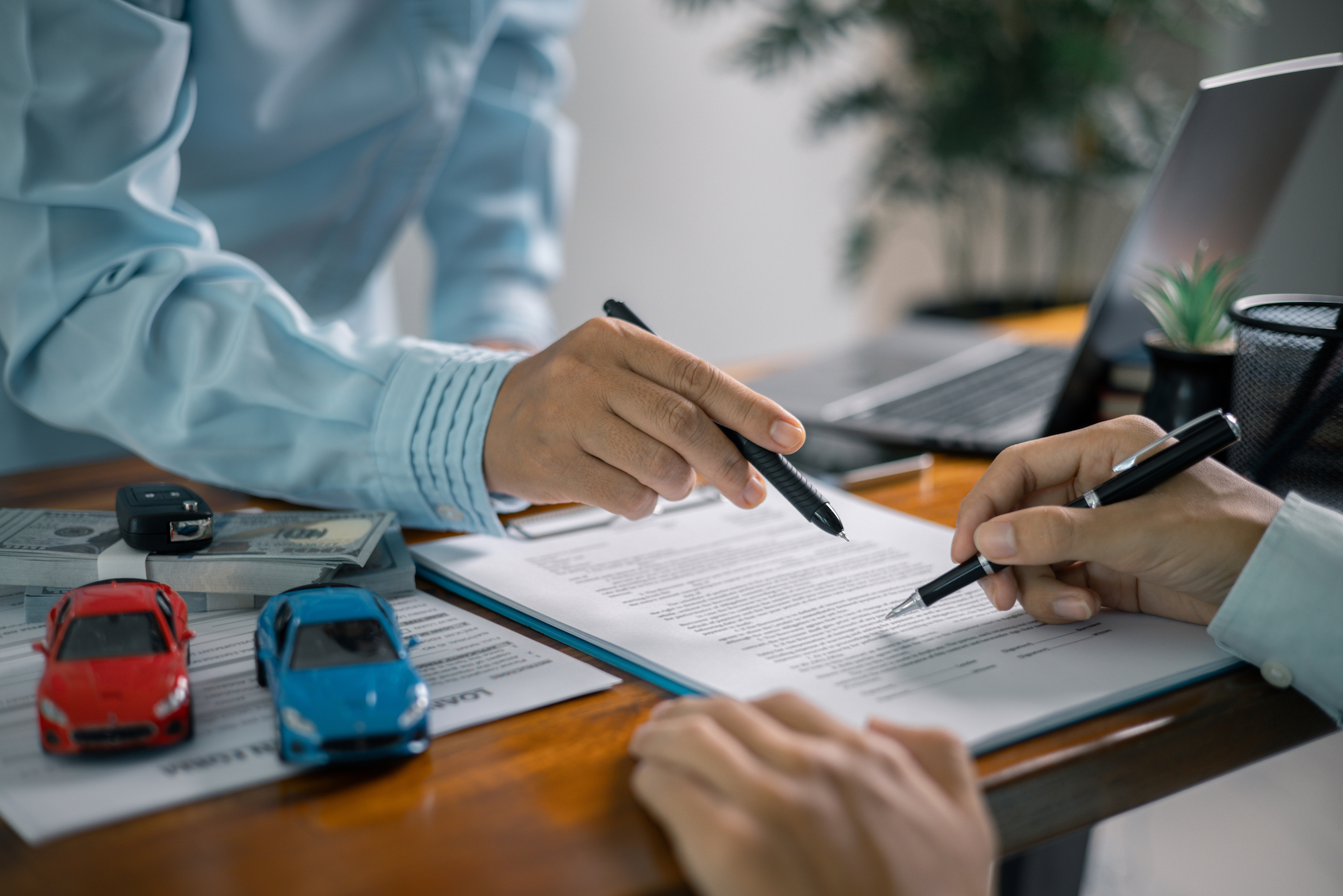 The image depicts two people signing a car purchase or leasing agreement, with toy cars, car keys, and financial documents on the table.