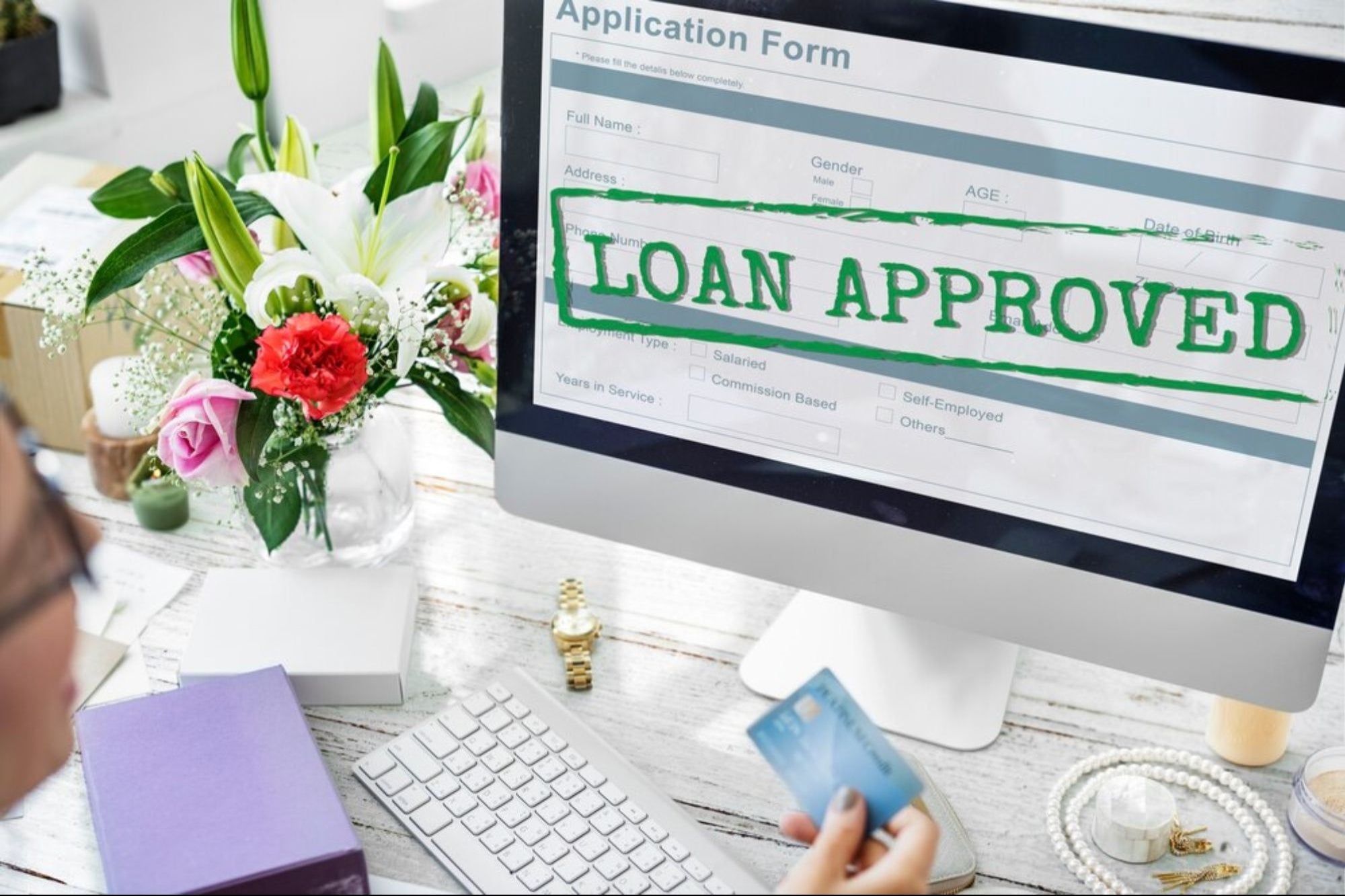 The image shows a person holding a credit card in front of a computer screen displaying an "LOAN APPROVED" message, with a desk decorated with flowers, jewelry, and office supplies.
