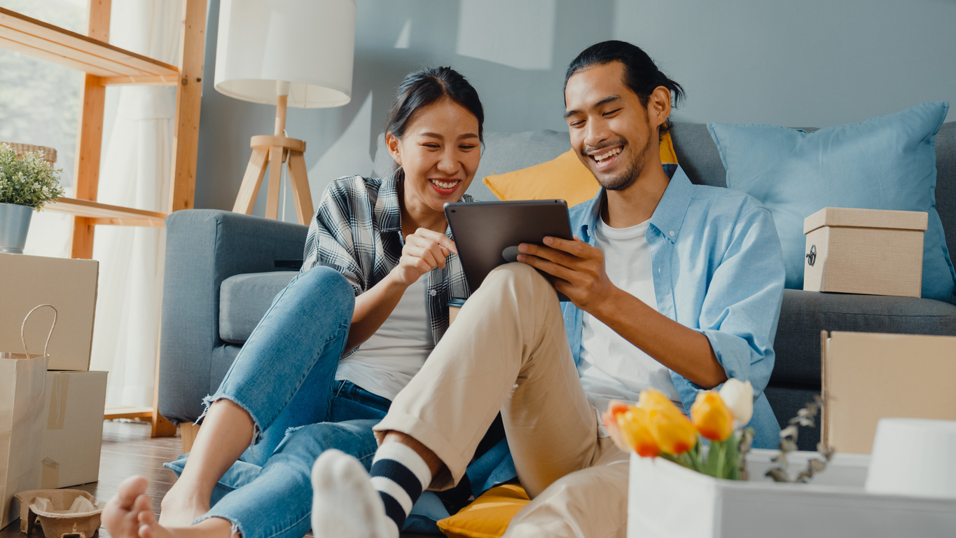 The picture shows a happy couple sitting on the floor, looking at a tablet together, surrounded by moving boxes, indicating they are likely in the process of moving into a new home.