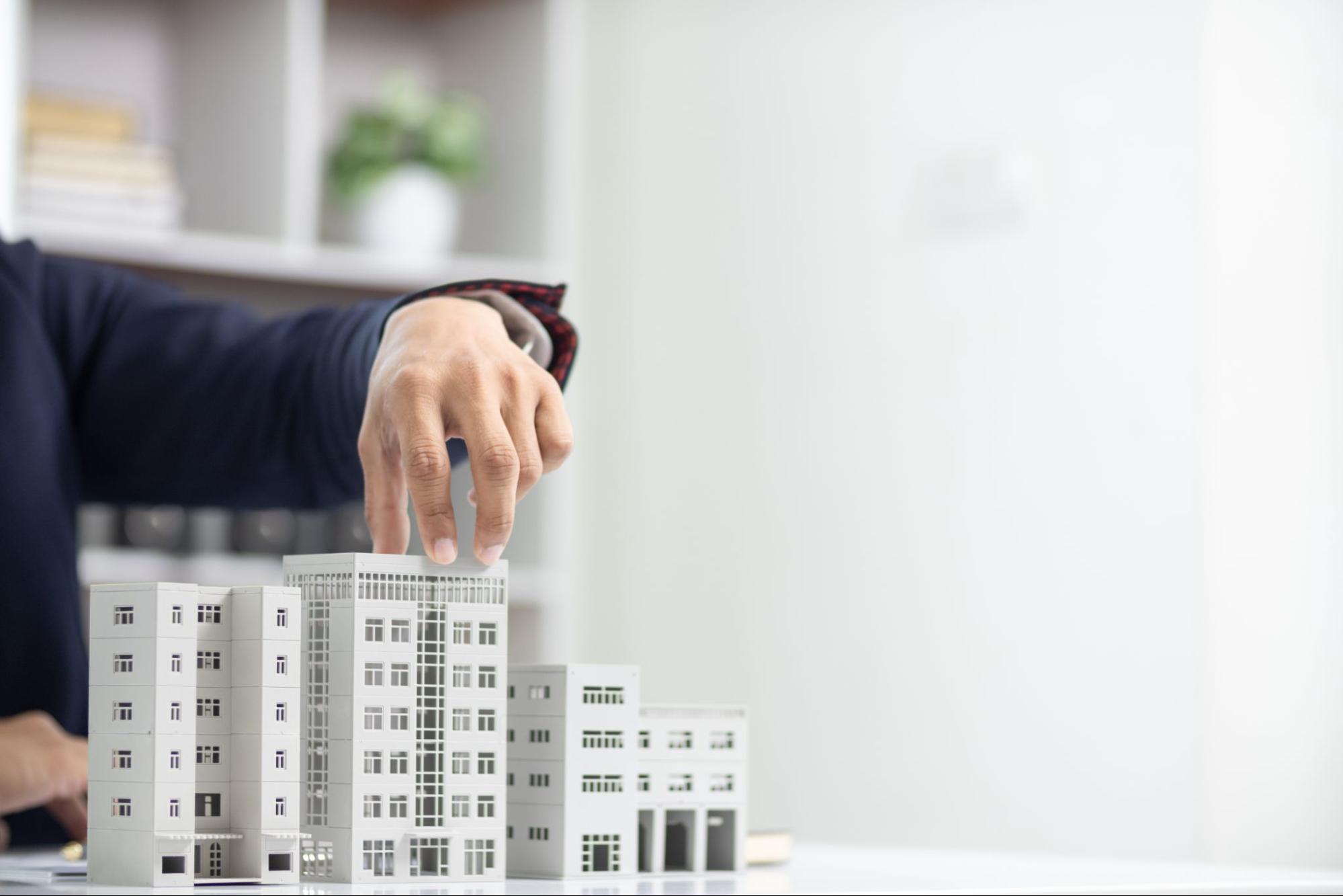 The image shows a person’s hand adjusting a miniature model of a building, likely representing real estate or property development.