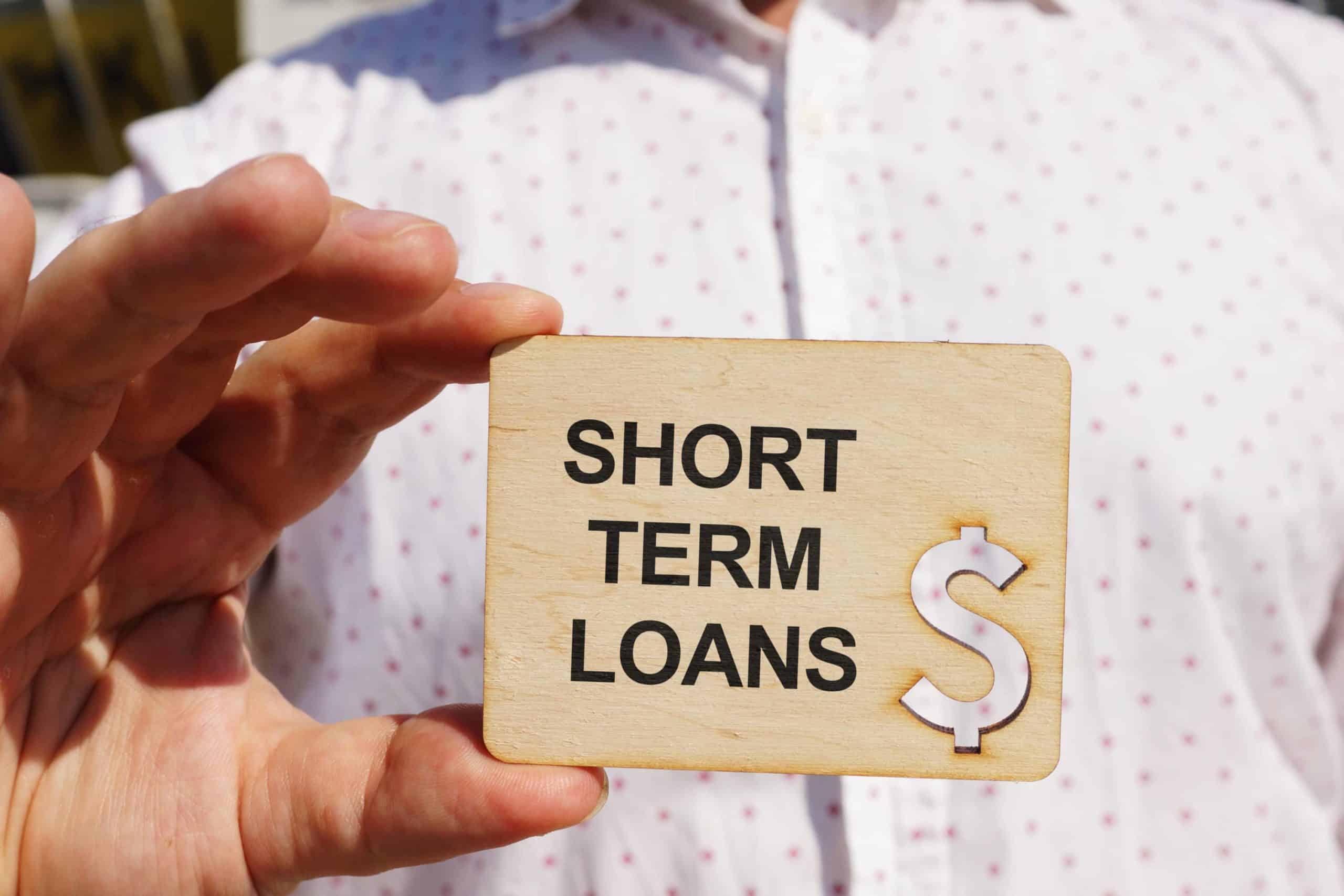 The image shows a person holding a wooden sign that reads "SHORT TERM LOANS" with a dollar sign, indicating a focus on short-term borrowing options.