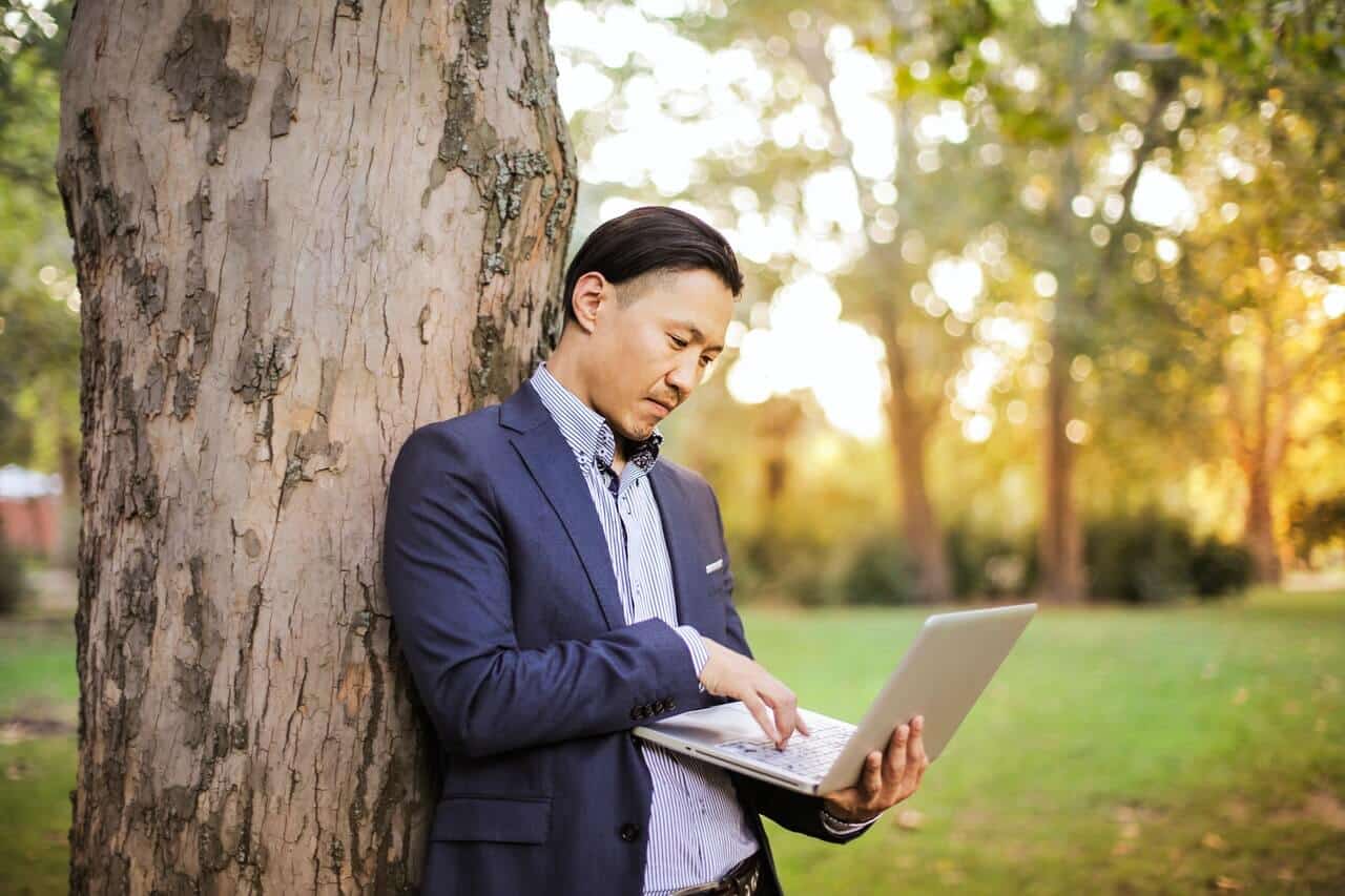 A man in a suit using a laptop outdoors, leaning against a tree in a park during sunset.