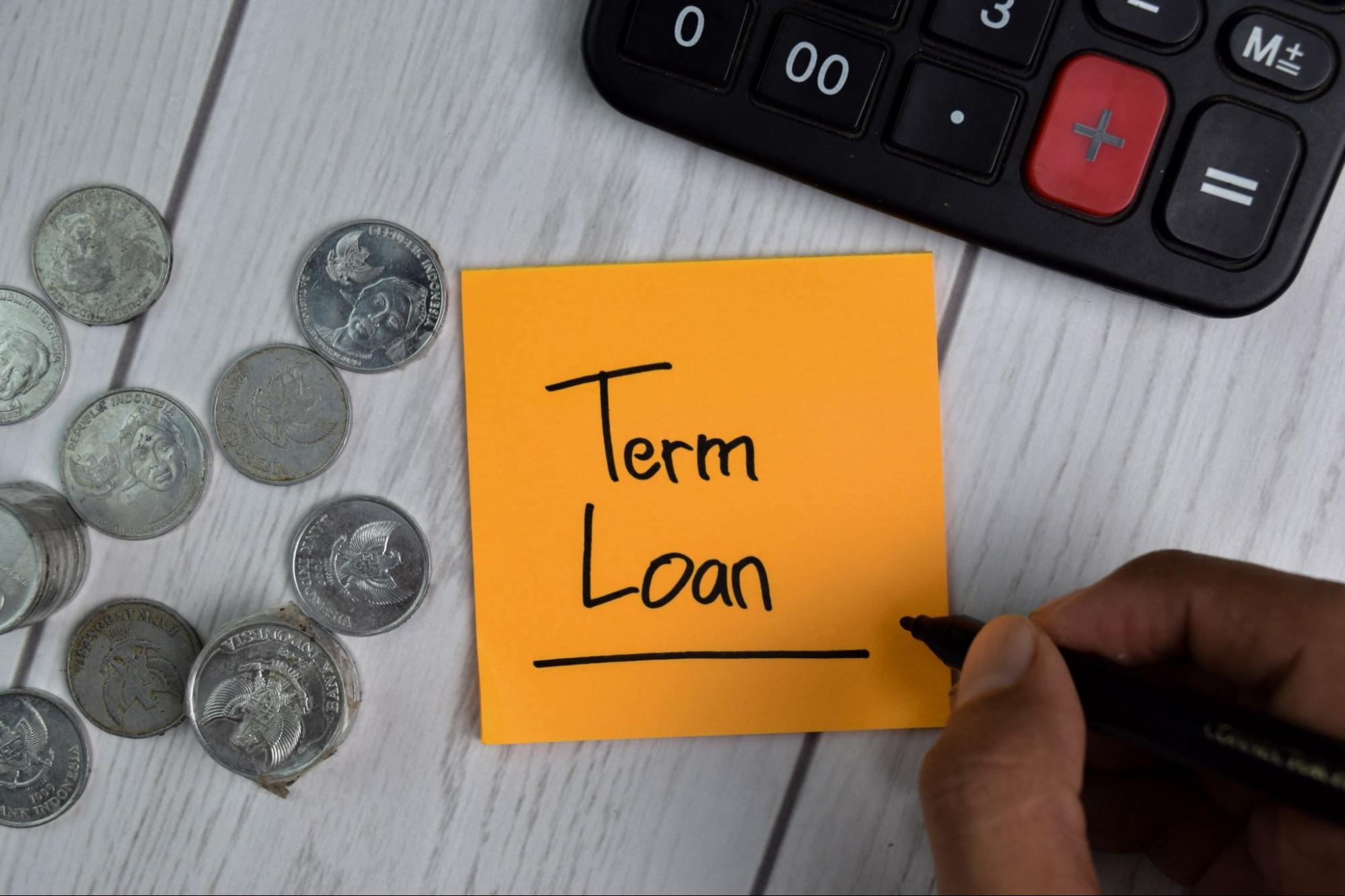 A person writing "Term Loan" on an orange sticky note, surrounded by coins, a calculator, and a white wooden surface.
