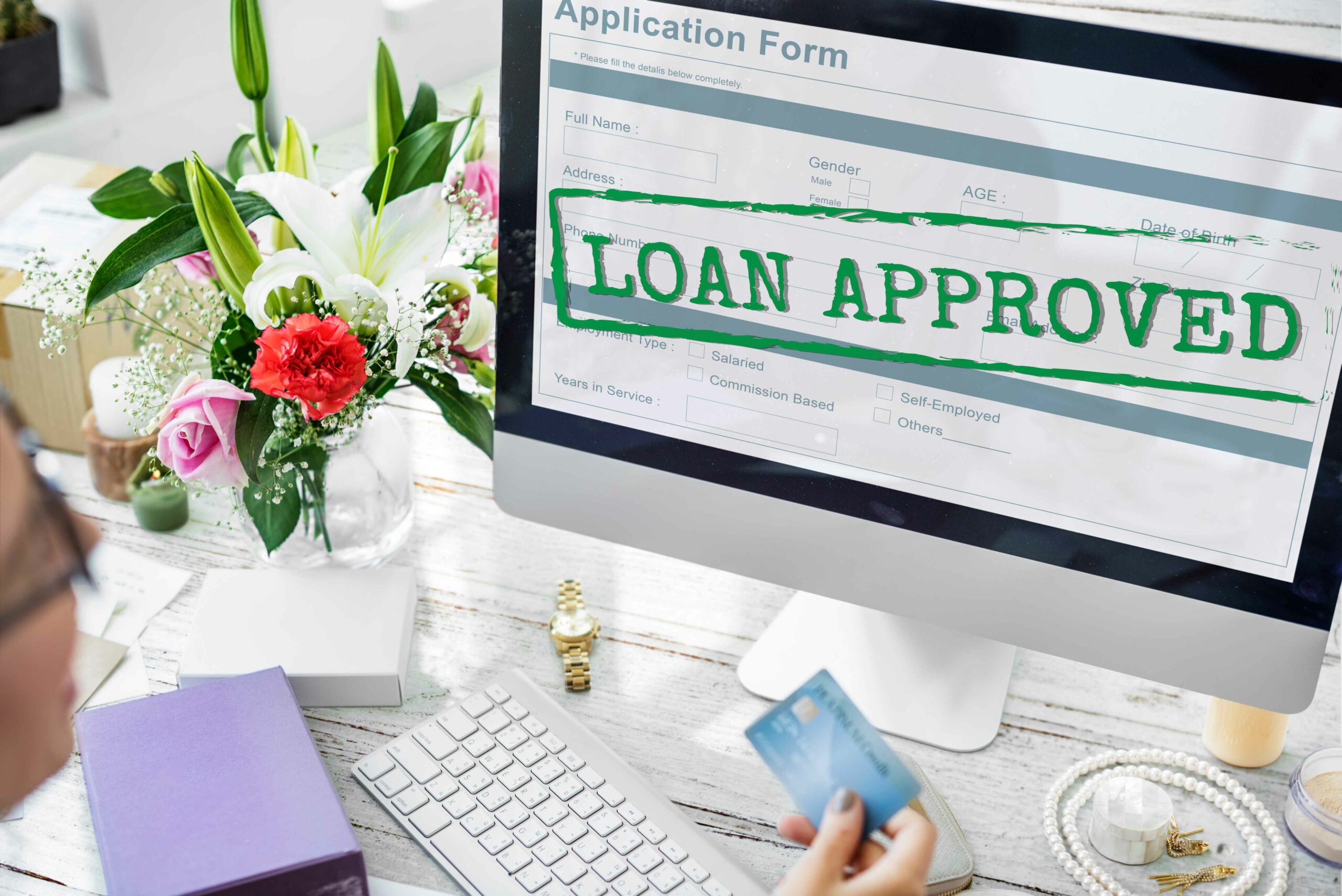 The image shows a person holding a credit card in front of a computer screen displaying an approved loan application.