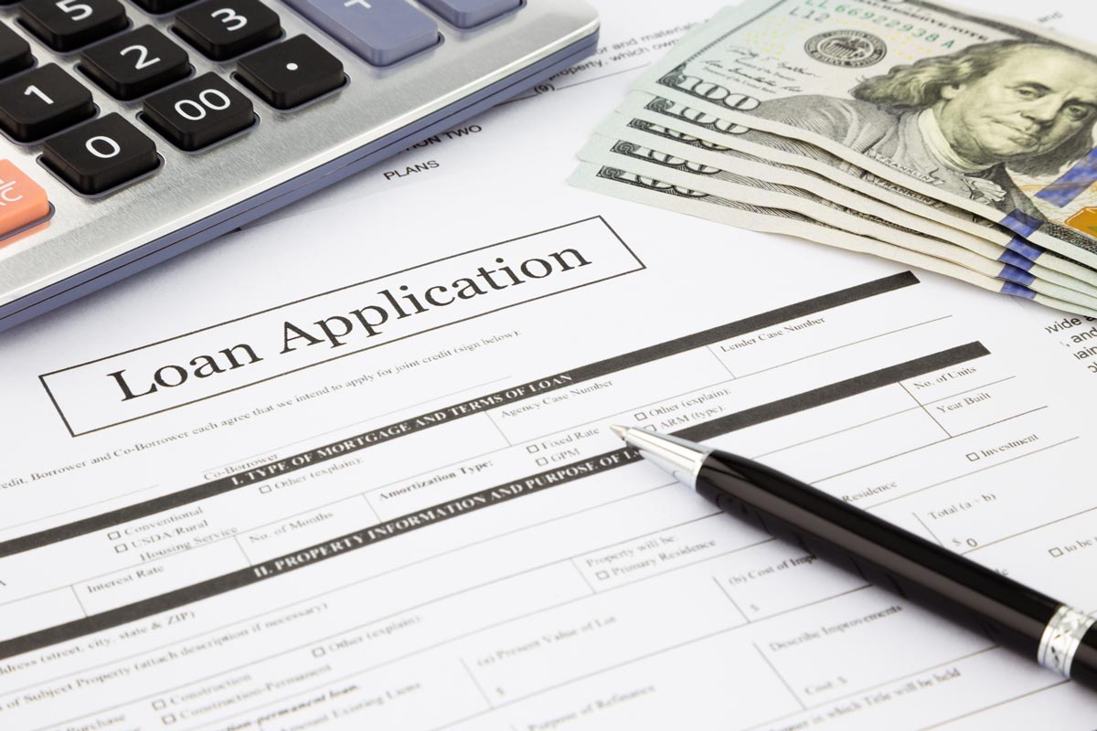 The picture depicts a loan application form alongside a pen, cash, and a calculator, suggesting the process of applying for a loan or financial planning.