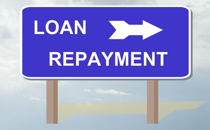 A large blue road sign with white text and an arrow pointing to the right. The sign reads "LOAN REPAYMENT." The sign is mounted on two wooden posts, and the background features a partly cloudy sky.