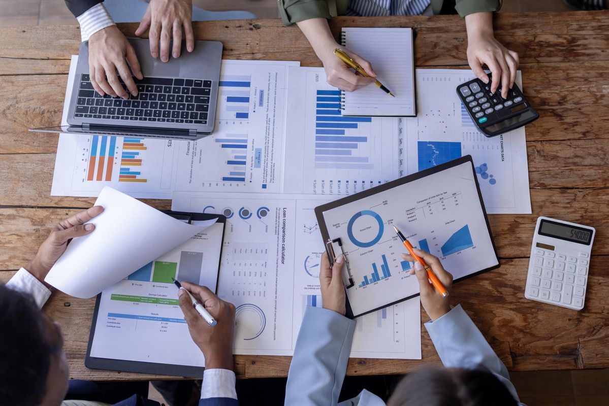 A top-down view of a business meeting with multiple people analyzing financial documents and charts spread out on a wooden table. The individuals are using laptops, tablets, notebooks, pens, a calculator, and printed graphs with various blue and orange ba