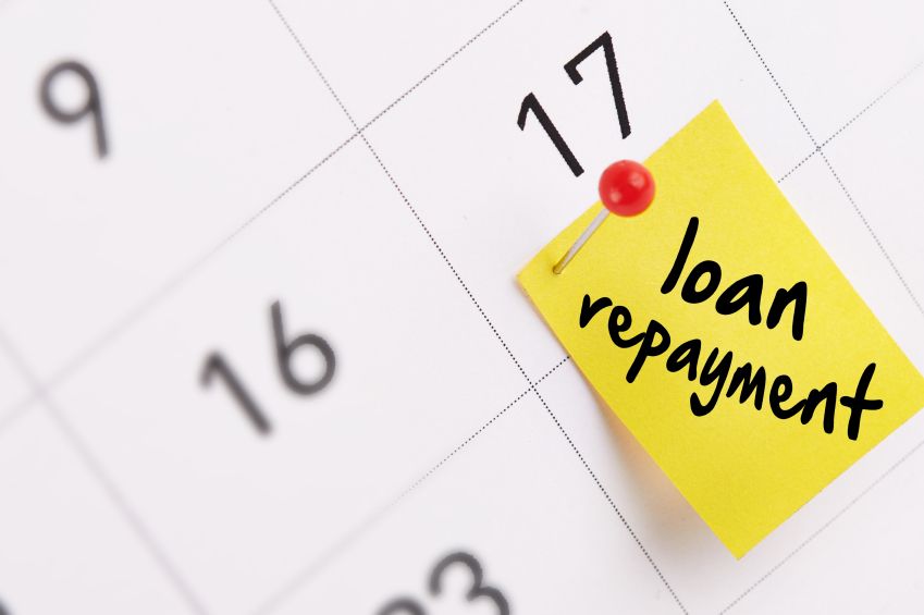 A close-up of a calendar with a yellow sticky note pinned to the date of the 17th. The sticky note has "loan repayment" written on it in bold black letters. The calendar shows the dates 16 and 17, with the note indicating an important deadline for loan re