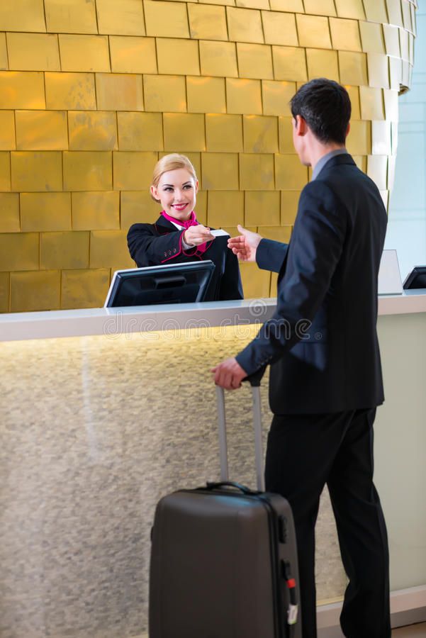 A hotel guest is happily checking in at the front desk, handing over his credit card while holding a rolling suitcase.