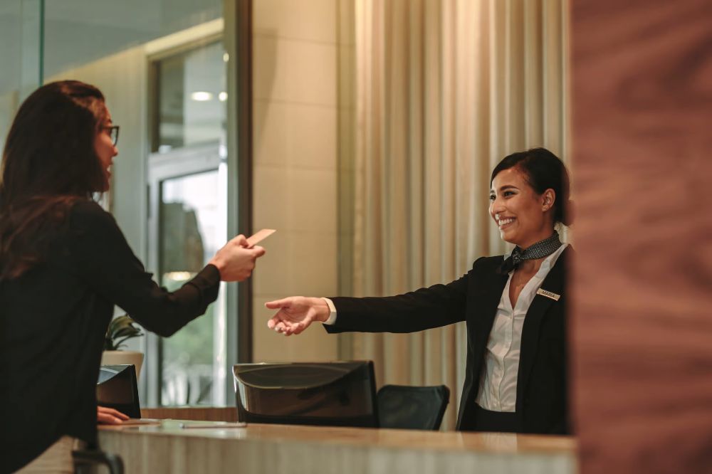 The image shows a cheerful hotel receptionist handing a key or card to a guest at the front desk.