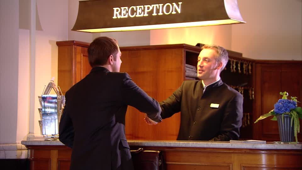The image depicts a guest greeting a hotel receptionist at the reception desk.