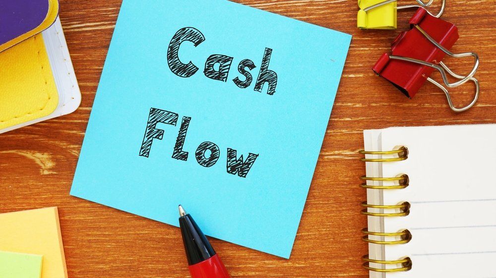 The image depicts a blue sticky note with the words "Cash Flow" written on it, surrounded by office supplies on a wooden desk.