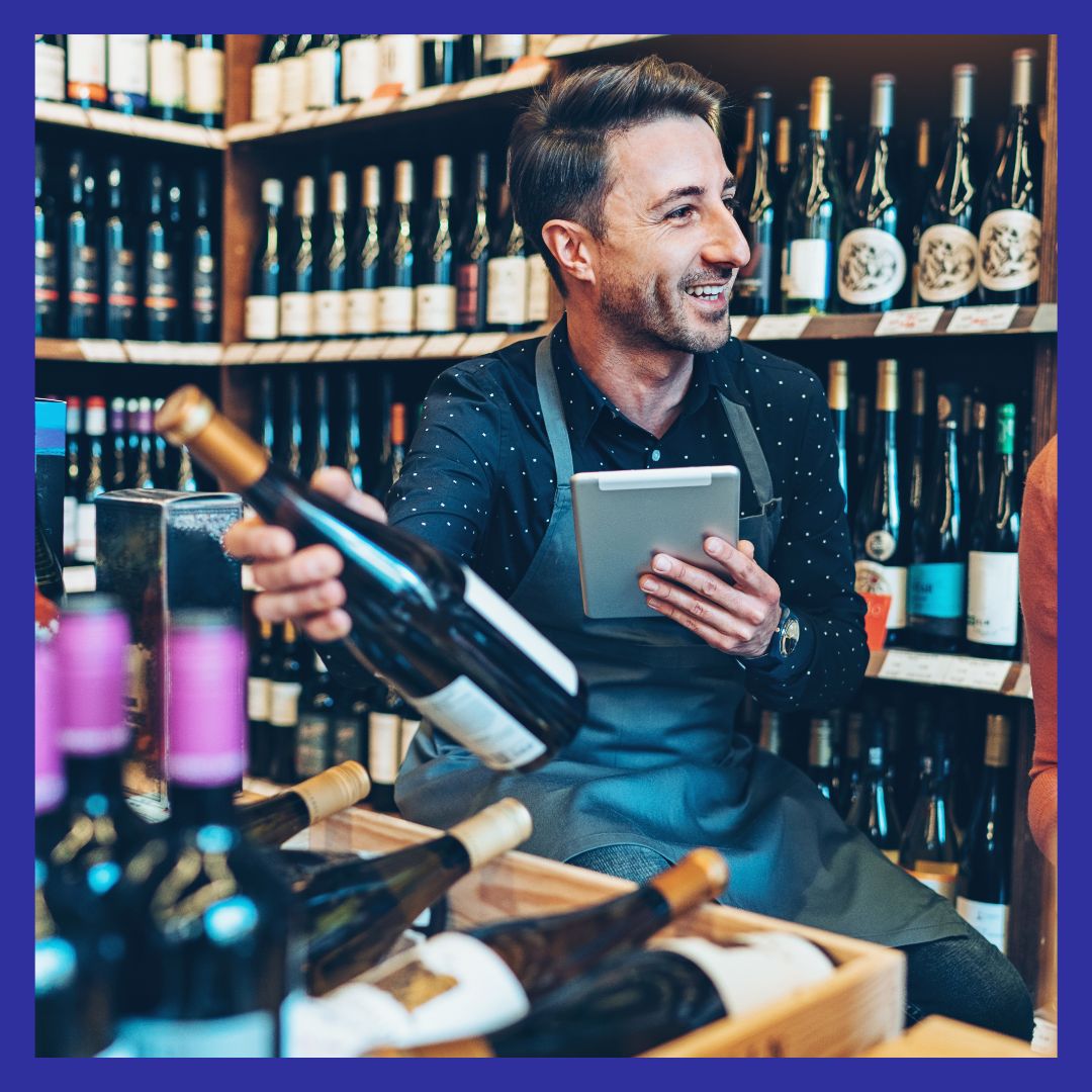 A cheerful man working as a wine store clerk, holding a wine bottle and a tablet amid shelves of wine bottles.