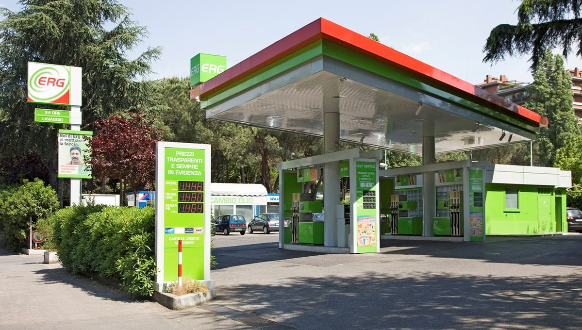 A green and white ERG gas station with self-service fuel pumps set amidst trees and parked cars on a sunny day.