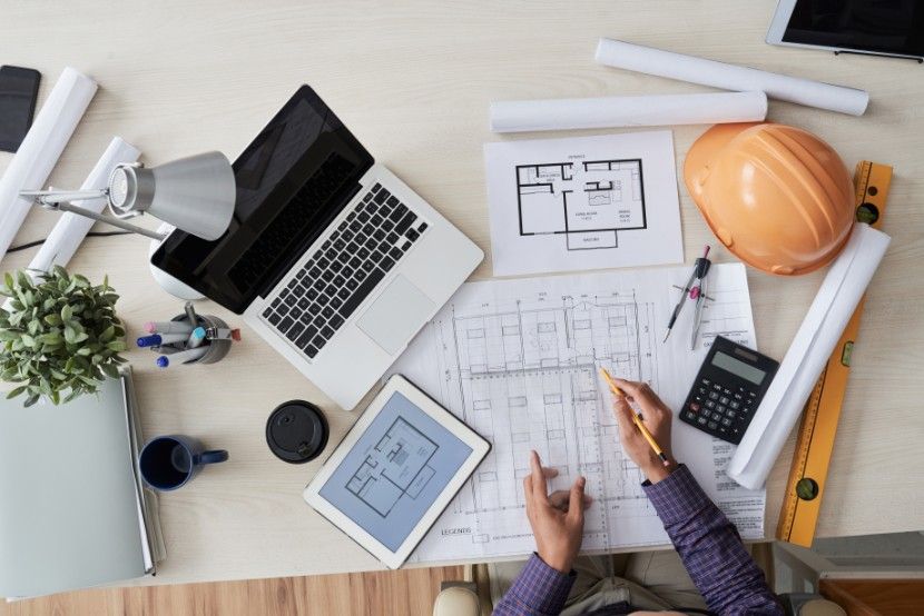 A top-down view of an architect's workspace featuring blueprints, a laptop, a tablet with building plans, a hard hat, and various drafting tools.
