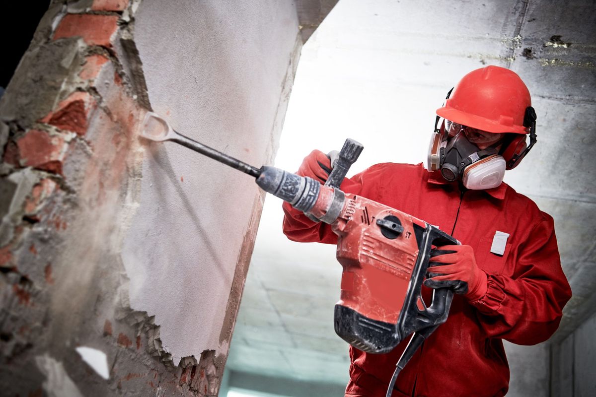 A construction worker wearing safety gear using a jackhammer to break through a wall.