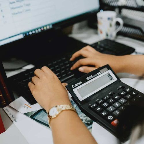 A person working at a desk using a computer keyboard, calculator, and smartphone, with a coffee mug in the background.