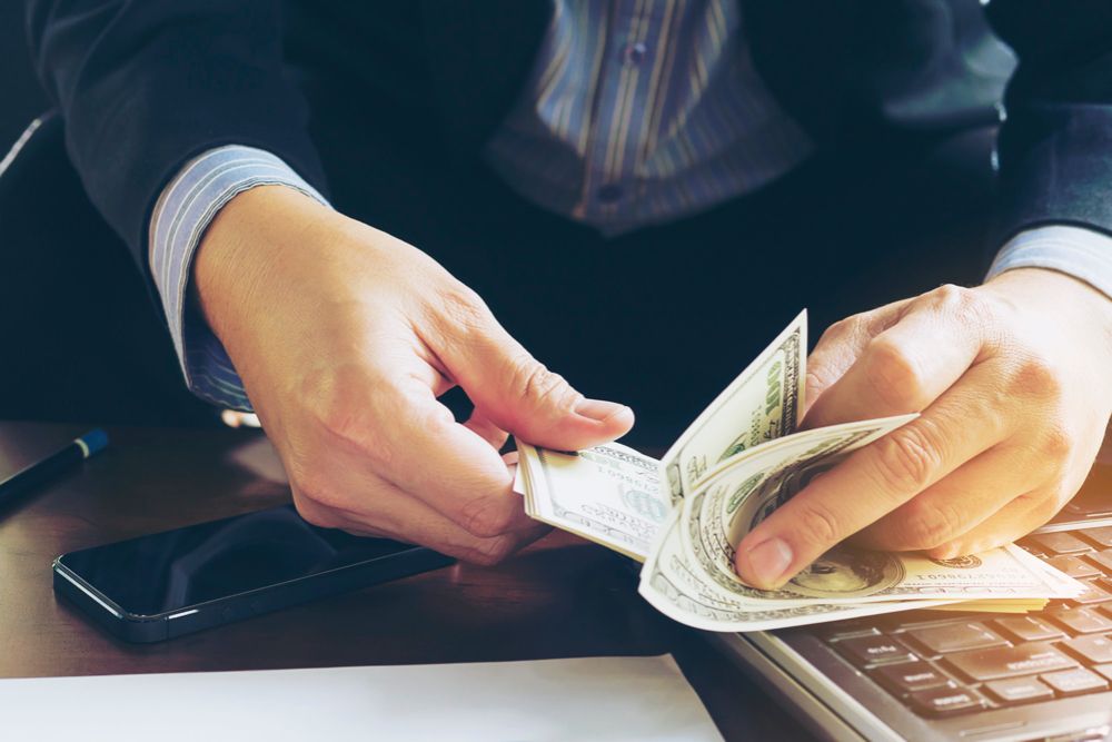 Hands counting cash beside a laptop and smartphone, representing business funding or cash flow management.