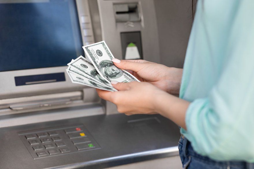 A person withdrawing cash from an ATM while holding U.S. dollar bills in front of the machine.