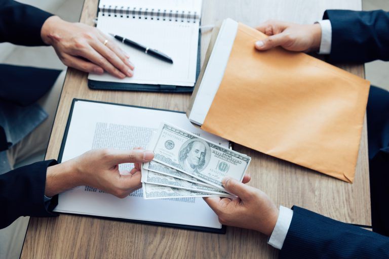 Two business professionals exchanging U.S. dollar cash across a desk with documents and a folder, symbolizing a financial agreement or business funding transaction.