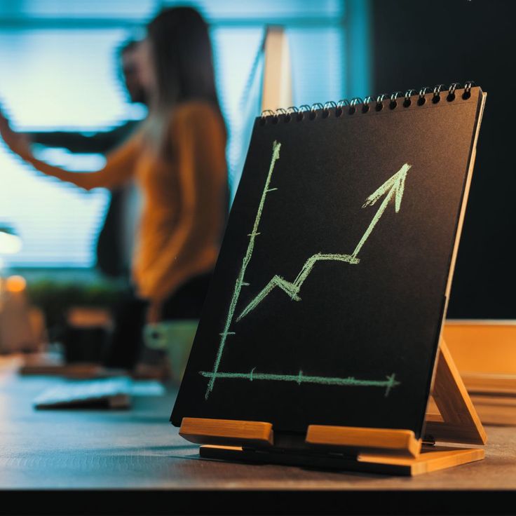 A small chalkboard on a desk displays an upward-trending line graph with an arrow, symbolizing business or financial growth, with blurred professionals discussing in the background.