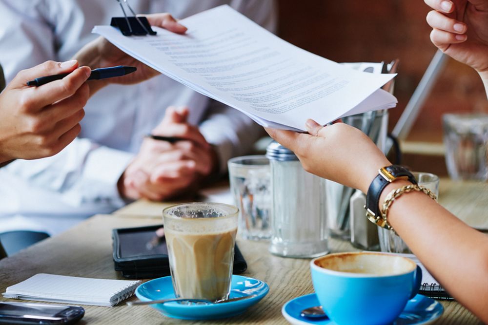 Business professionals reviewing and discussing printed documents over coffee at a table, symbolizing contract negotiation, financial consultation, or merchant cash advance agreement discussions.