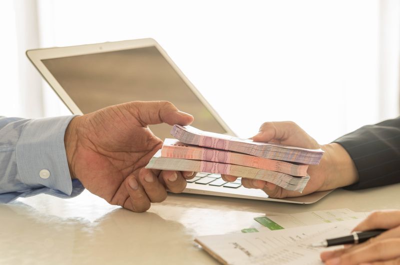 Two people exchanging a stack of cash across a desk with a laptop and paperwork, representing a business loan, merchant cash advance, or financial agreement.