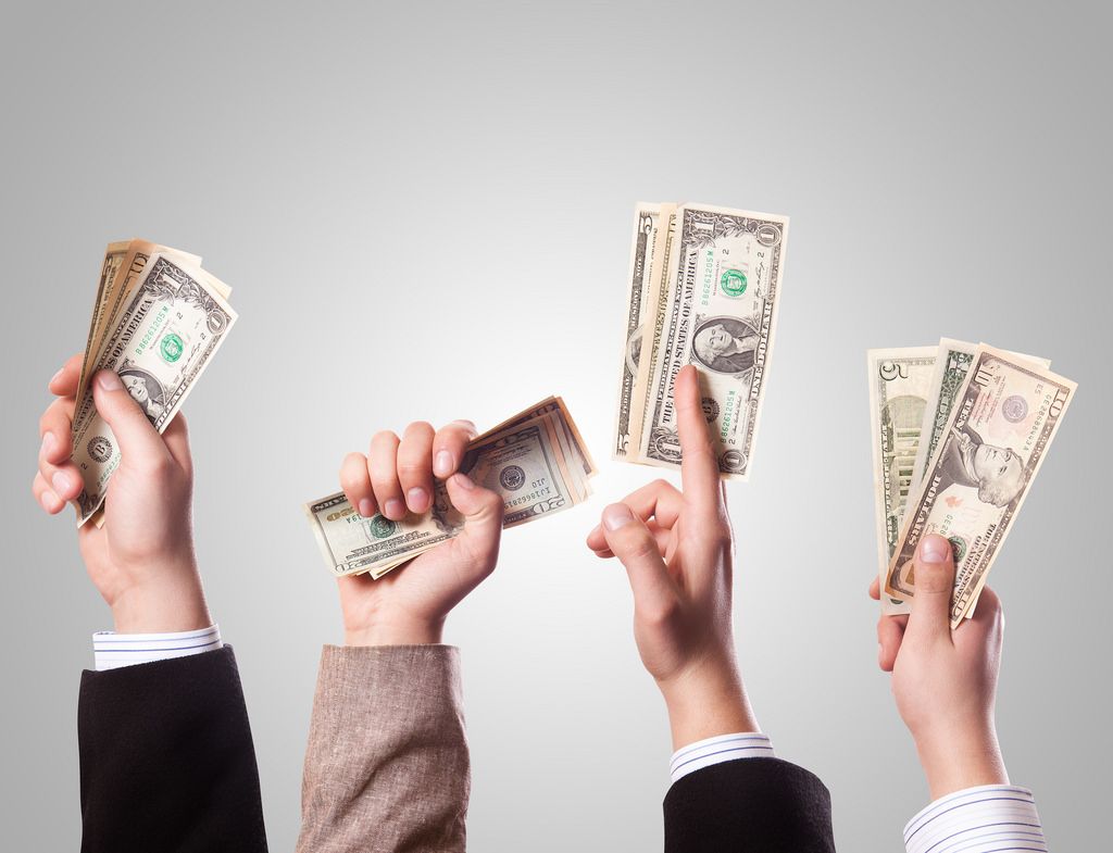 Multiple hands raised holding stacks of U.S. dollar bills against a plain background, symbolizing payments, income, cash flow, or financial transactions.