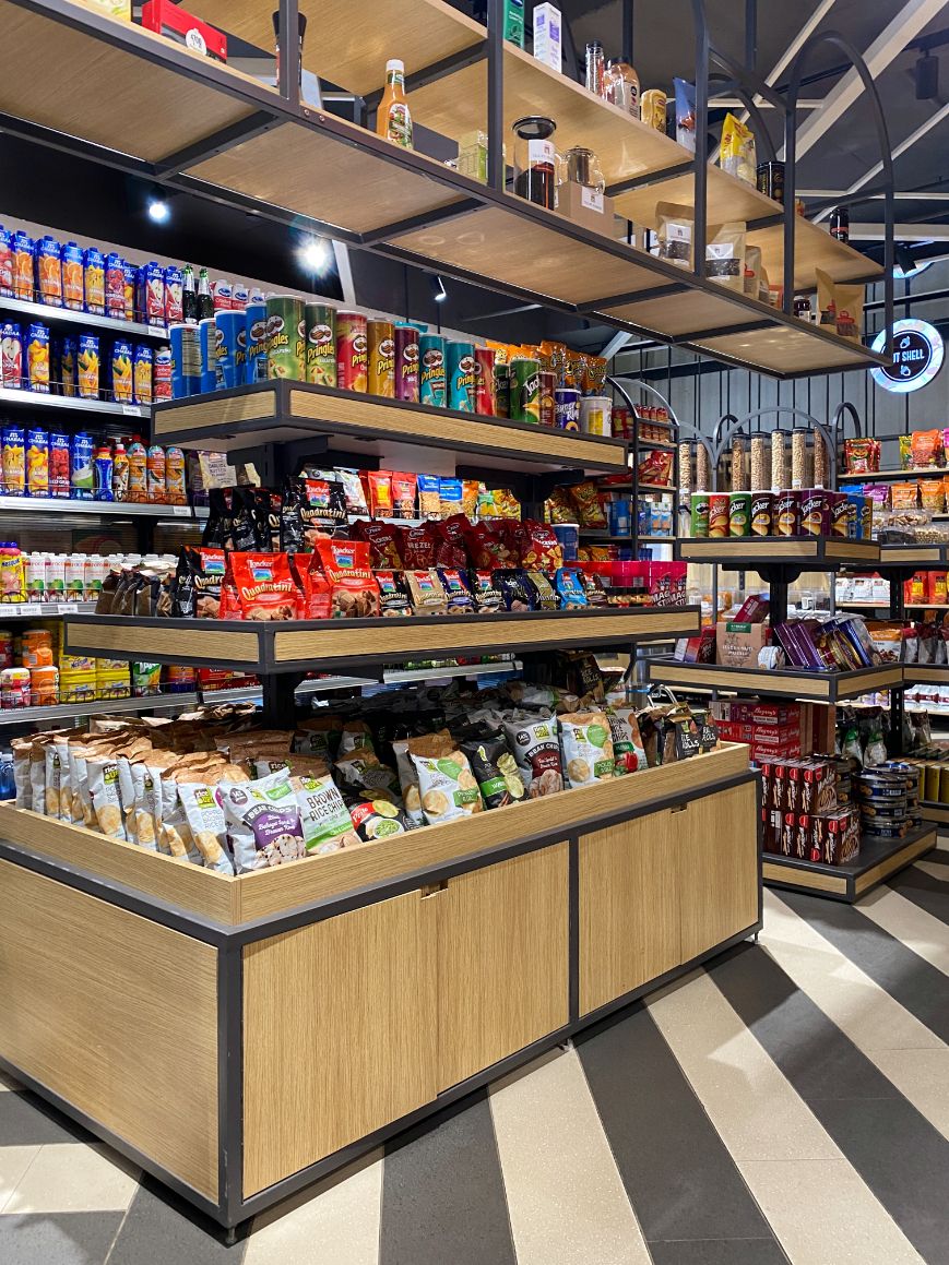 A modern grocery store interior with neatly arranged shelves and wooden display units filled with packaged snacks, chips, beverages, and pantry items under bright retail lighting.