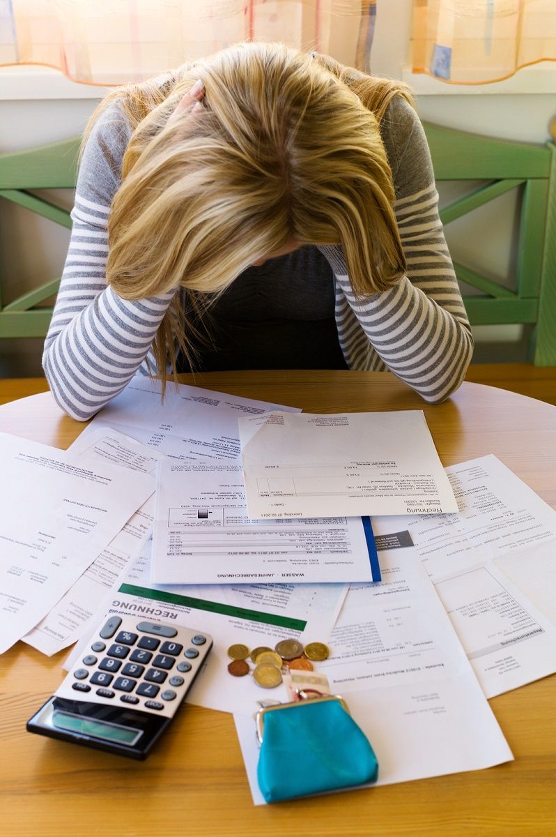 A person sitting at a table with their head in their hands, surrounded by bills, paperwork, a calculator, and loose coins, depicting financial stress and difficulty managing expenses.