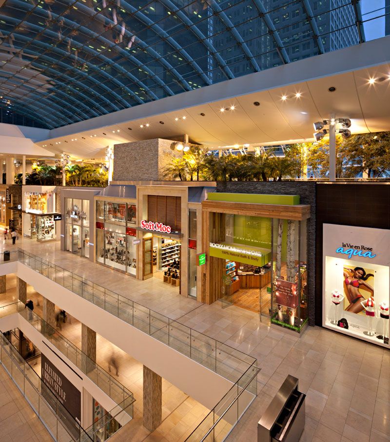 Interior of a modern shopping mall with multiple floors, glass railings, branded retail stores, indoor plants, and a large glass ceiling allowing natural light.